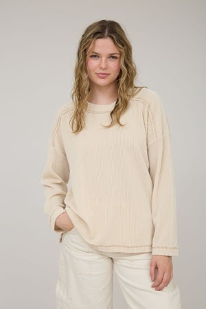 Woman wearing a beige sweater and white pants against a plain background