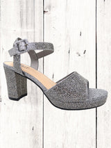 A black heel with a rhinestone design on the heel, featuring a one-strap design across the toe and an adjustable ankle strap on a white wooden surface.