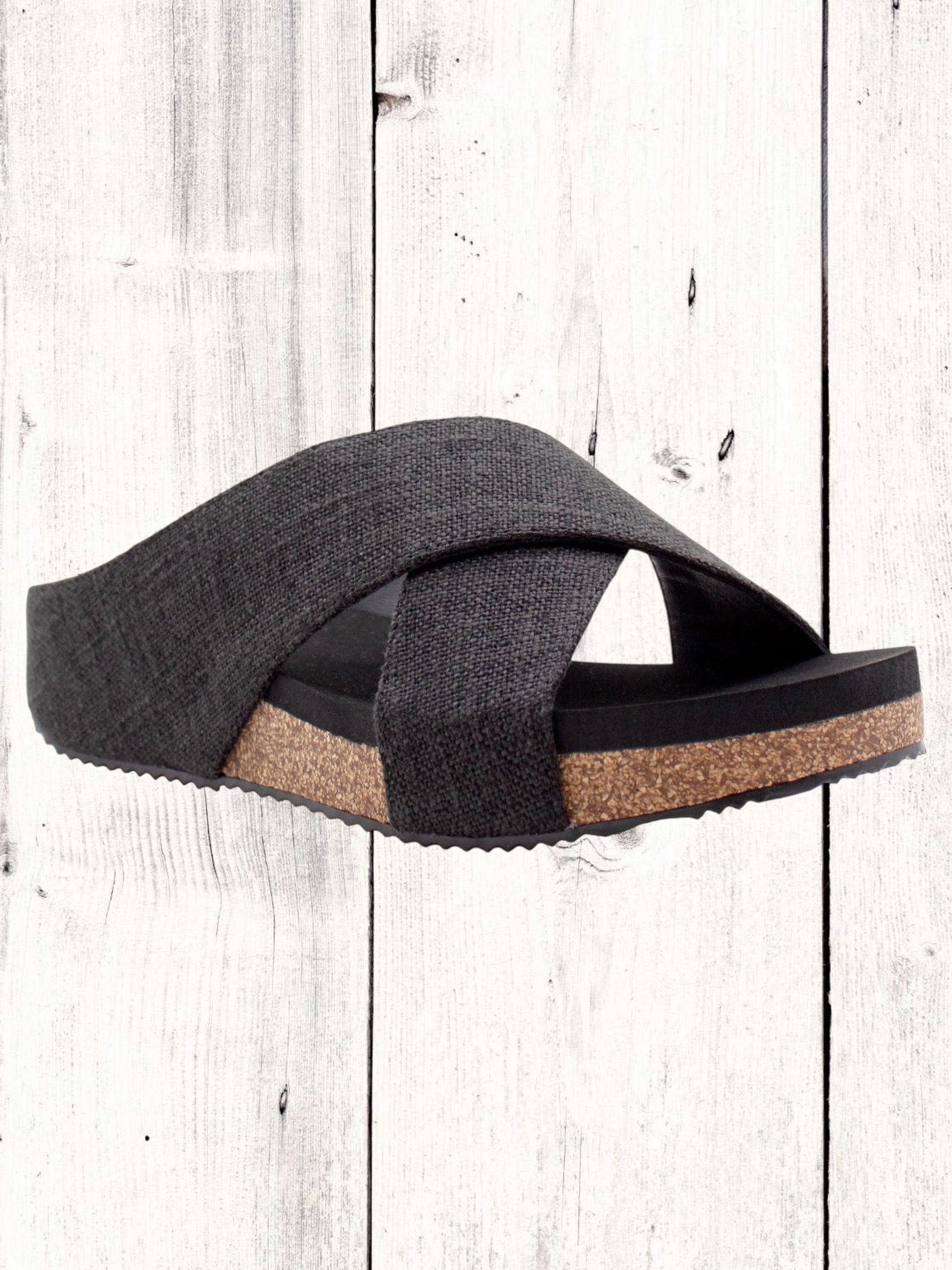 A pair of beige crisscross slide sandals with a low wedge heel, displayed against a wooden background.