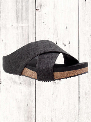 A pair of beige crisscross slide sandals with a low wedge heel, displayed against a wooden background.