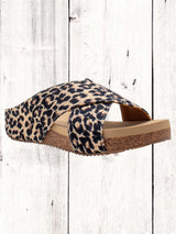 A pair of beige crisscross slide sandals with a low wedge heel, displayed against a wooden background.