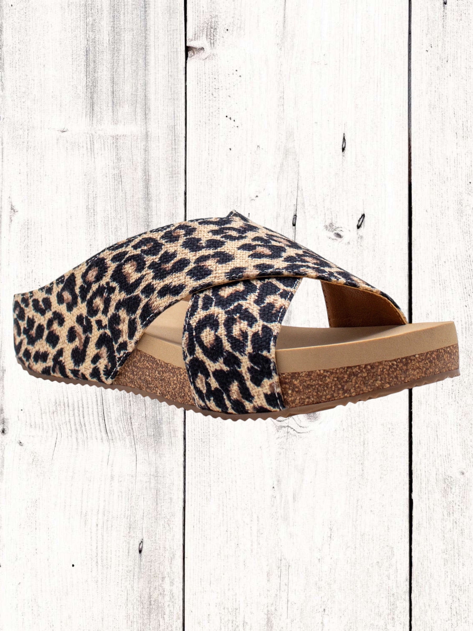A pair of beige crisscross slide sandals with a low wedge heel, displayed against a wooden background.