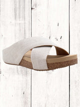 A pair of beige crisscross slide sandals with a low wedge heel, displayed against a wooden background.