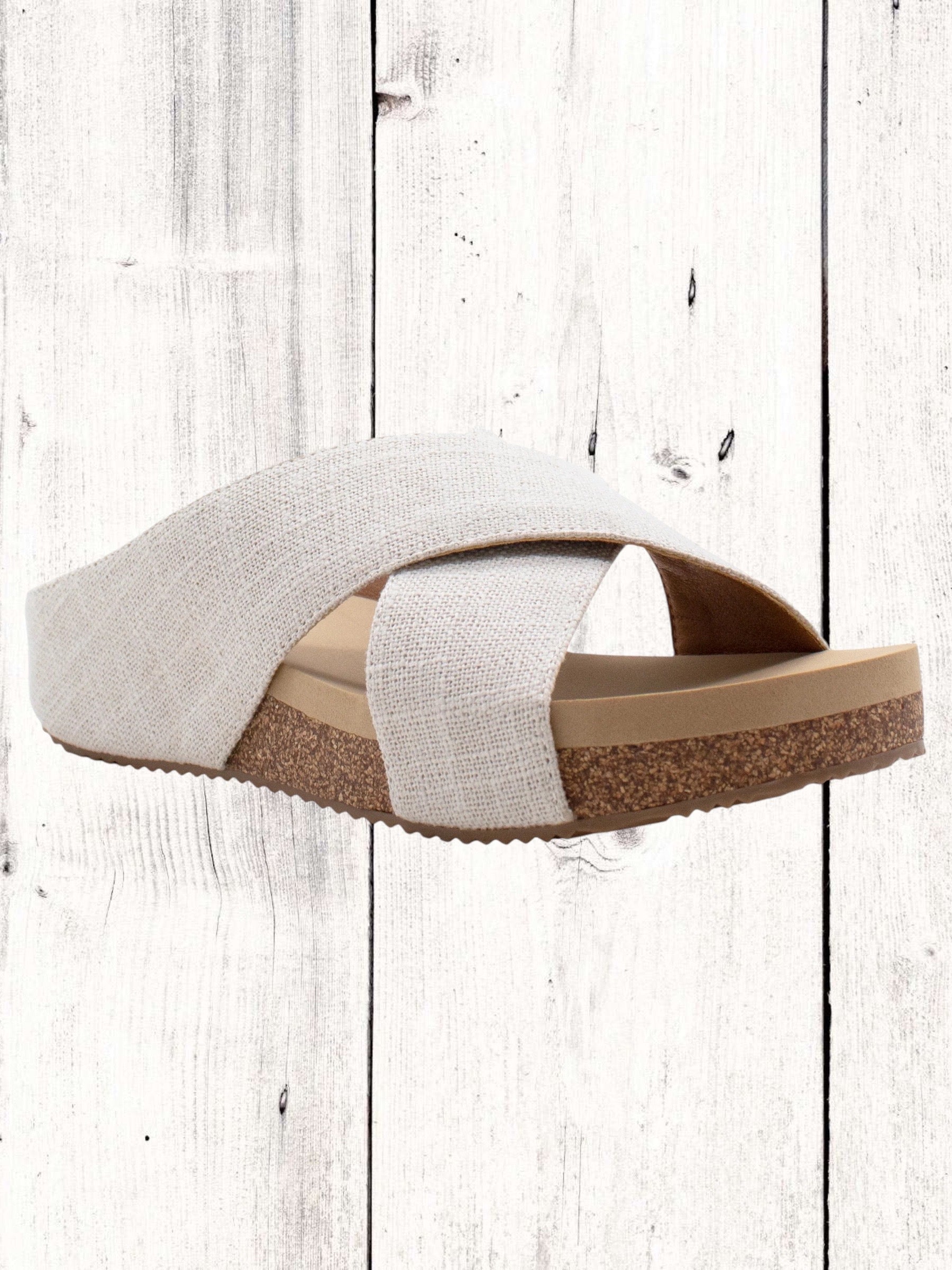 A pair of beige crisscross slide sandals with a low wedge heel, displayed against a wooden background.