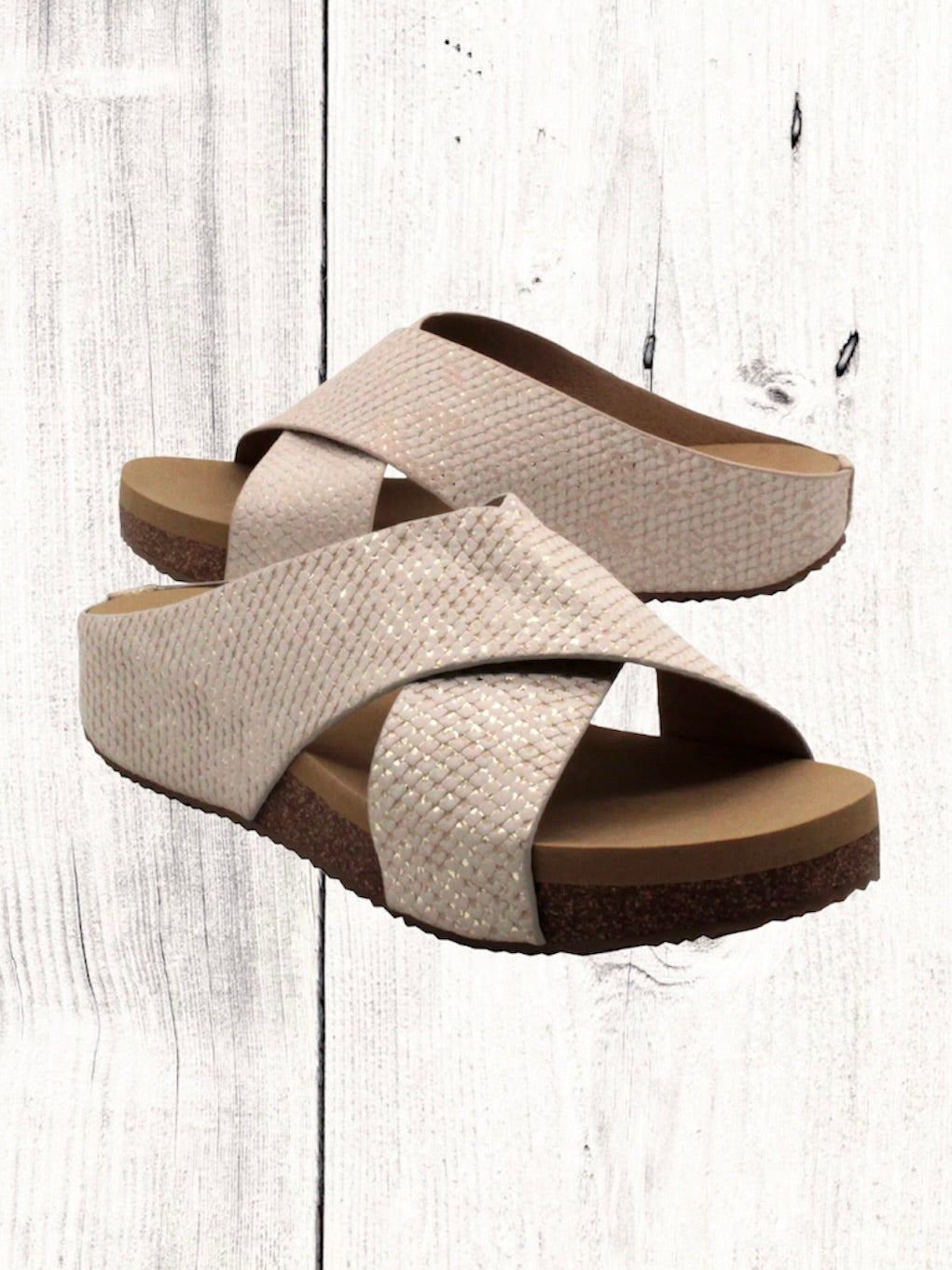 A pair of beige crisscross slide sandals with a low wedge heel, displayed against a wooden background.