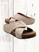 A pair of beige crisscross slide sandals with a low wedge heel, displayed against a wooden background.