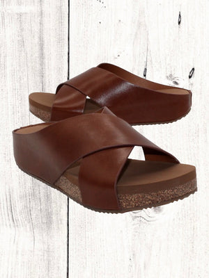 A pair of beige crisscross slide sandals with a low wedge heel, displayed against a wooden background.