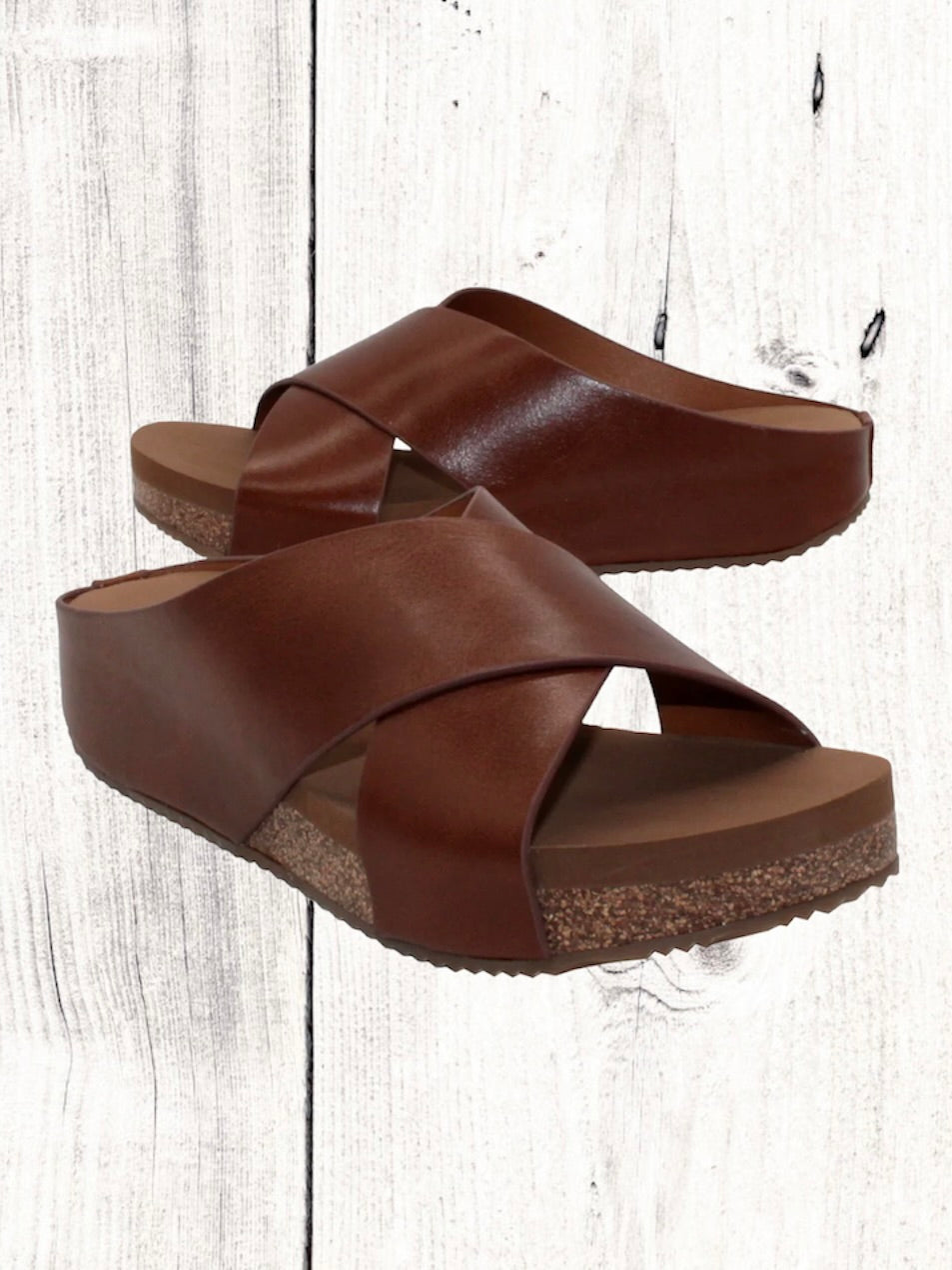 A pair of beige crisscross slide sandals with a low wedge heel, displayed against a wooden background.
