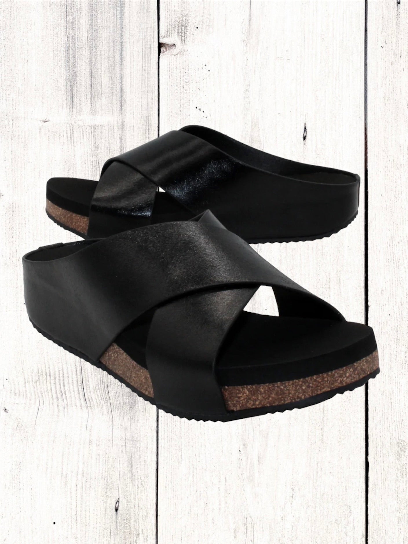 A pair of beige crisscross slide sandals with a low wedge heel, displayed against a wooden background.