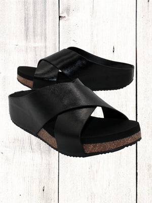 A pair of beige crisscross slide sandals with a low wedge heel, displayed against a wooden background.